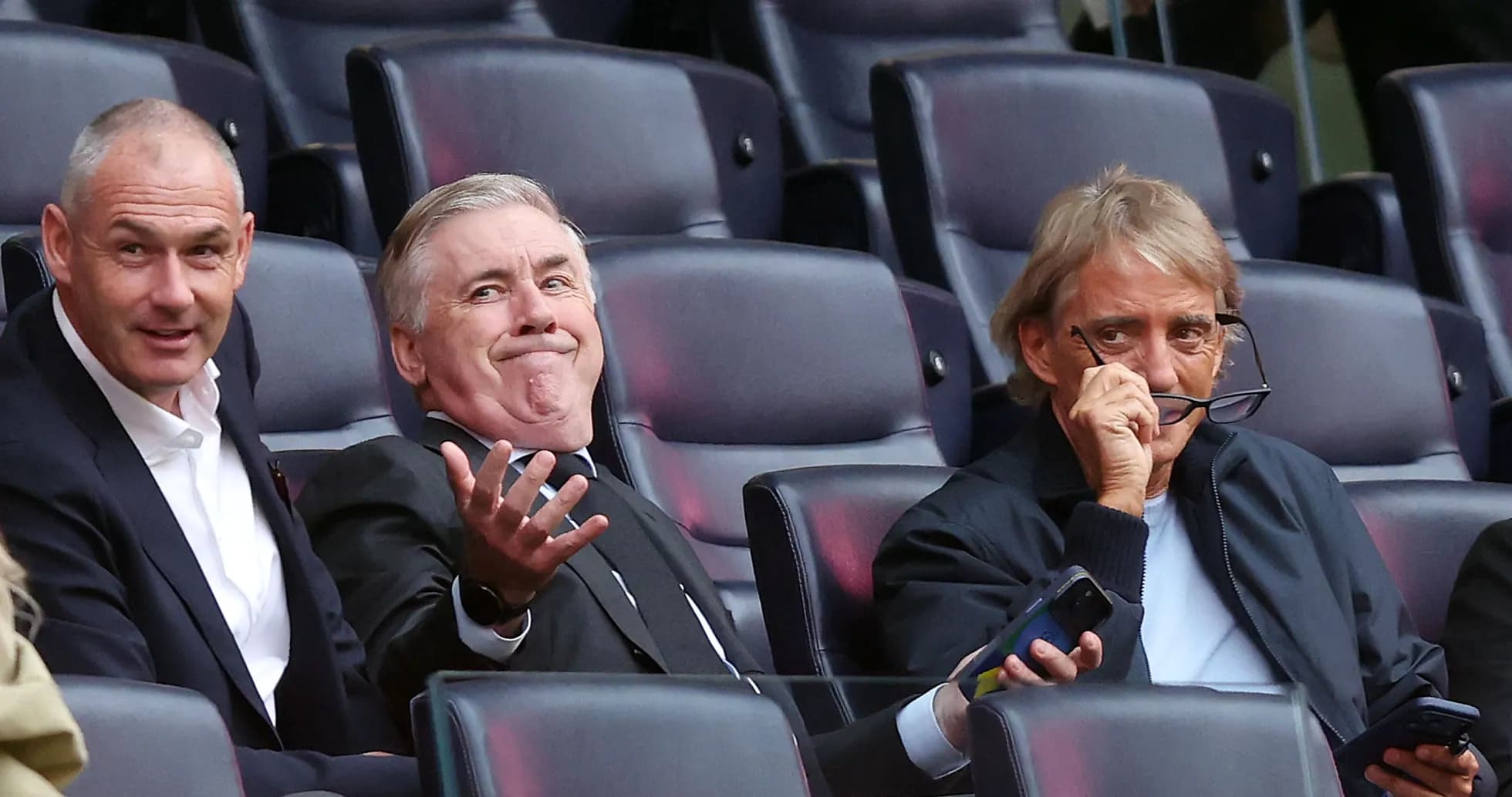 Three men seated in stadium, engaging in conversation